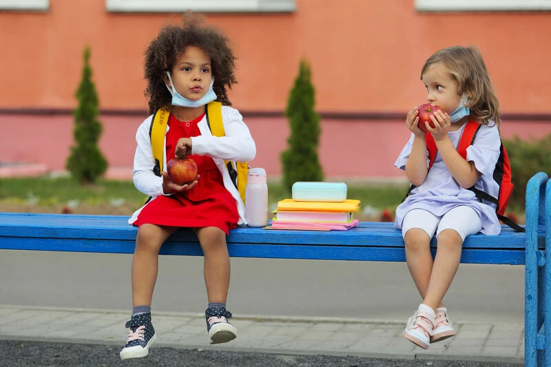 Two children sitting on a bench with their school lunches in between them